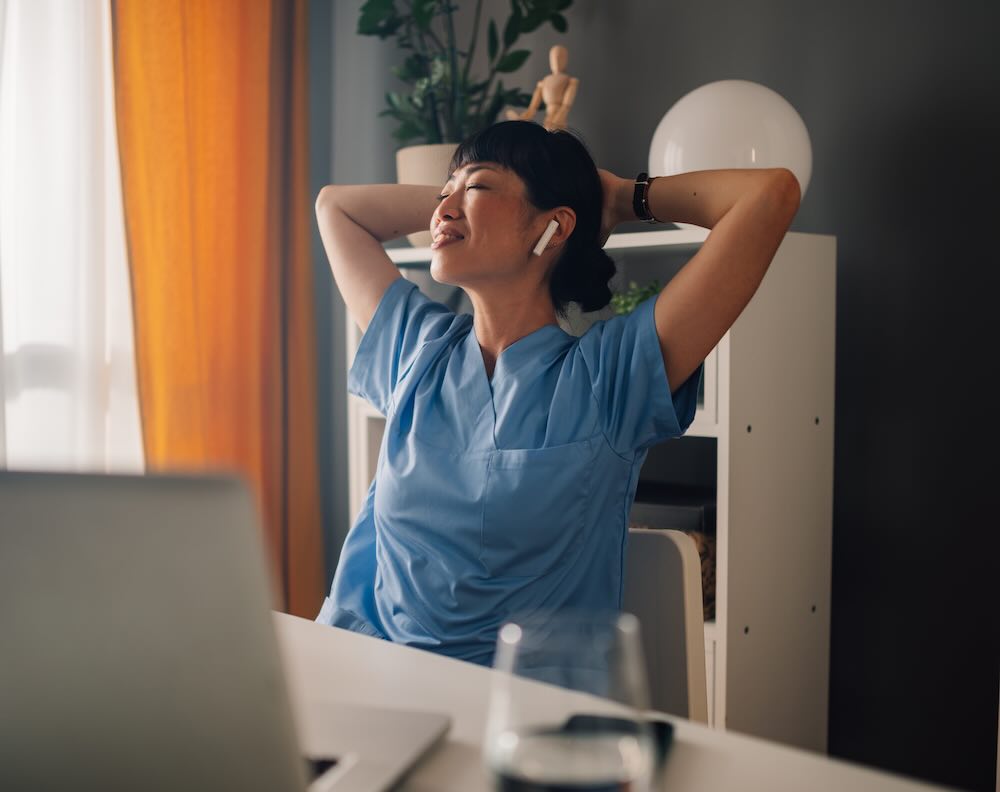 Relaxed asian woman nurse in blue scrubs stretching while sitting at desk 