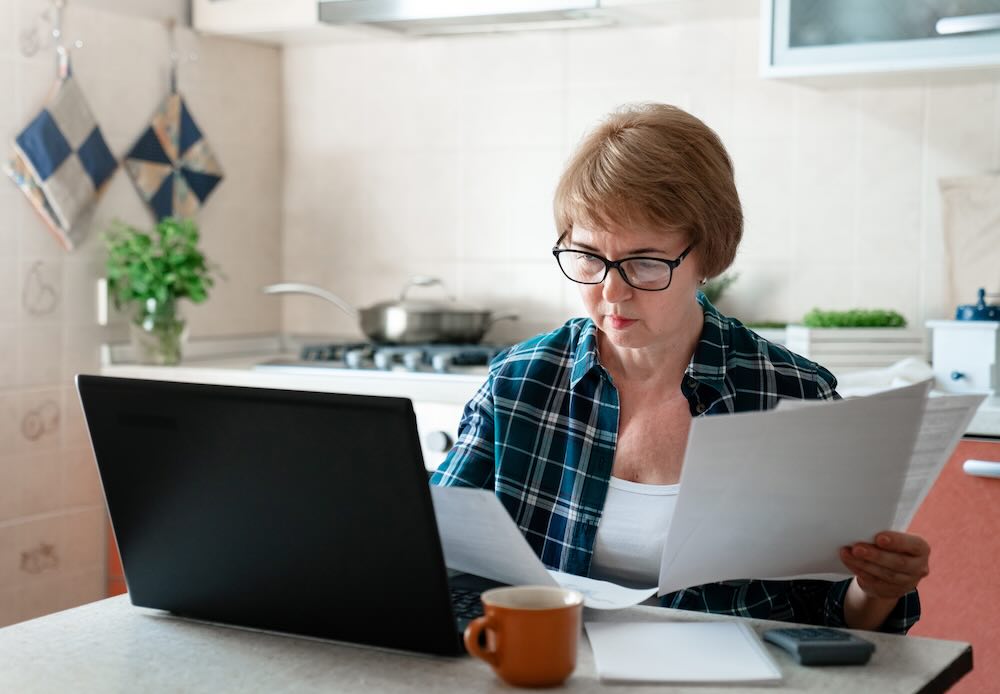 Senior woman working from home kitchen with laptop and documents